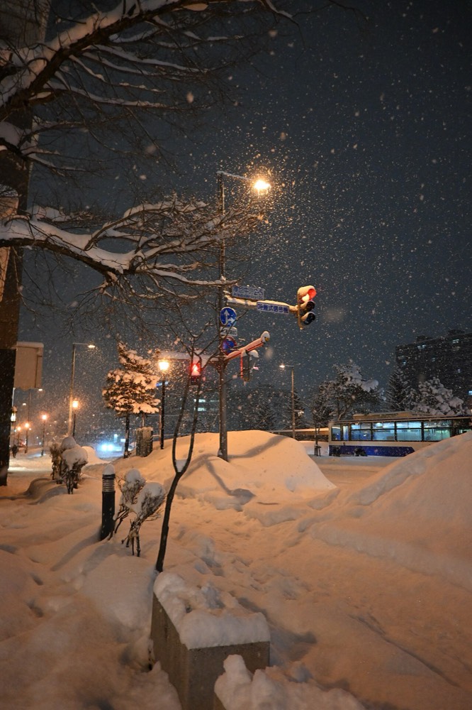 A snowing Sapporo street at night with thick snow piled along the roadside